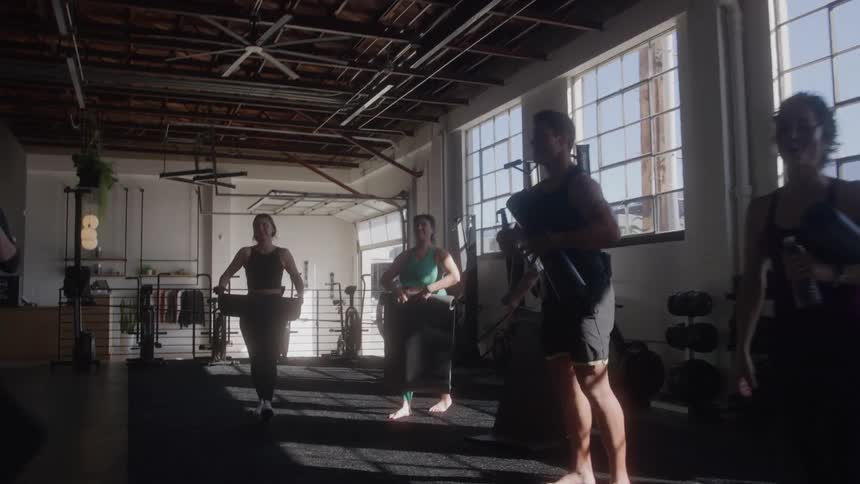 A group of people finish a workout and pack up mats in a sunlit gym