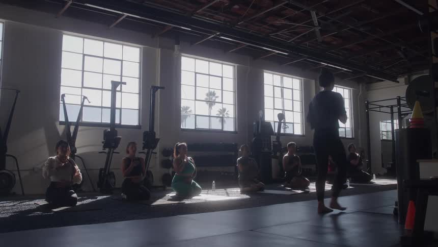 A large group of men and women are doing yoga on a mat in a gym with a coach walking in front of them and bright light shining through tall windows