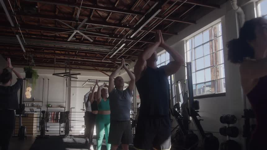 The video shows a group of people in a gym who are participating in a group exercise they begin with their arms outstretched then raise them above their heads before bringing their hands together in a prayer pose