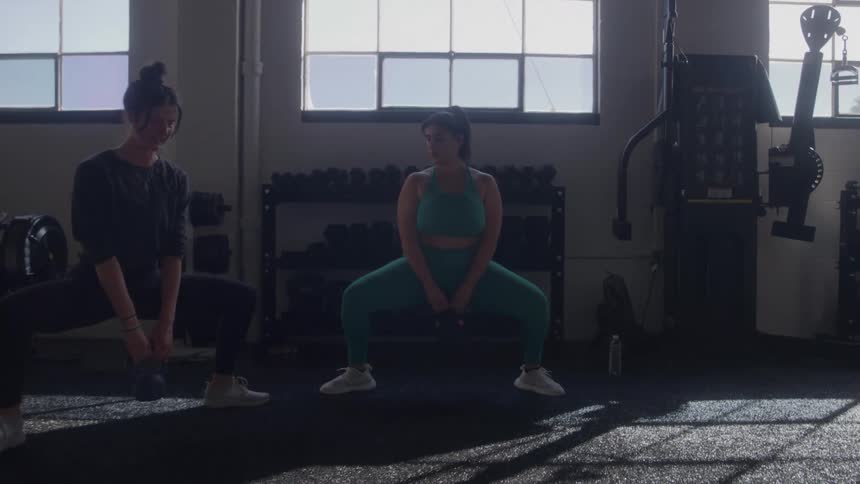 Two women perform weighted sumo squats in a bright gym faces showing concentrated effort