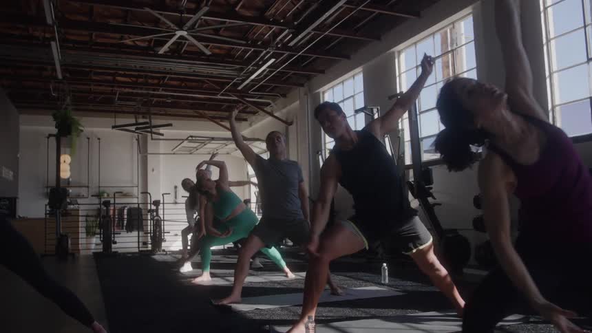A wide view shows a group of men and women of various ages doing a yoga pose with outstretched arms in a brightly lit gym with large windows