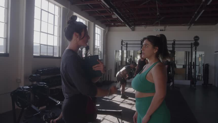 The video shows two women talking in a gym appearing to be a personal trainer and her client the trainer is demonstrating an exercise and giving instructions to the woman in the green sports bra