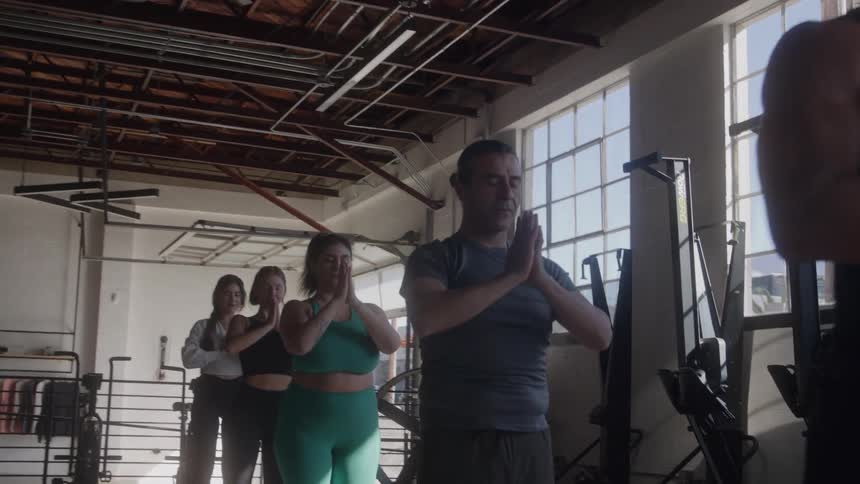 A diverse group of men and women are doing yoga together in a sunlit gym starting with their arms raised and then bringing their hands down to their chest