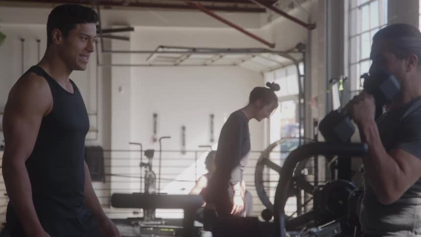 A fit male personal trainer is smiling while talking to his male client who is working out with dumbbells in a large gym