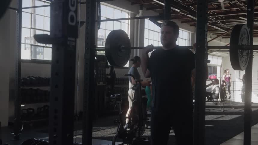 Man does squats while holding a weight bar at a busy gym during the daytime