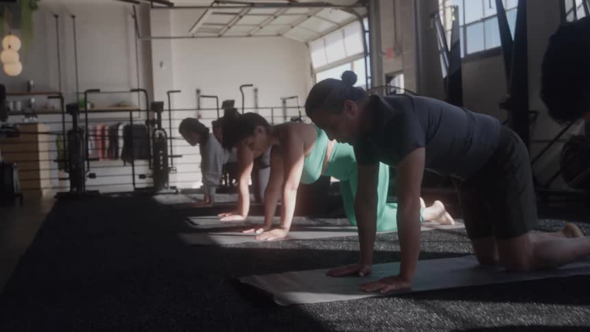 Group of males and females in a gym at midday bending and holding yoga poses on their mats