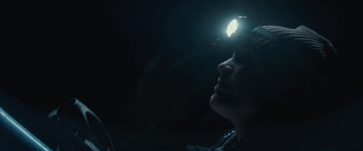 Man wearing a headlamp and cold weather outerwear uses a telescope before straightening to look up toward the sky at night