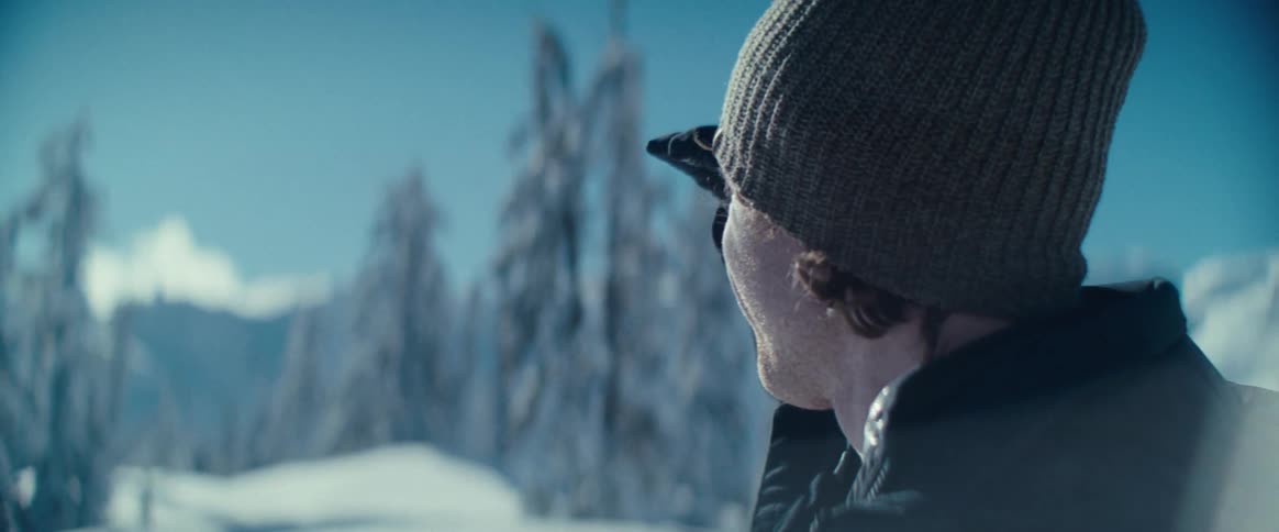 A man in a knit cap and jacket brings his hands up to his face while looking out over a bright snowy winter landscape with pine trees