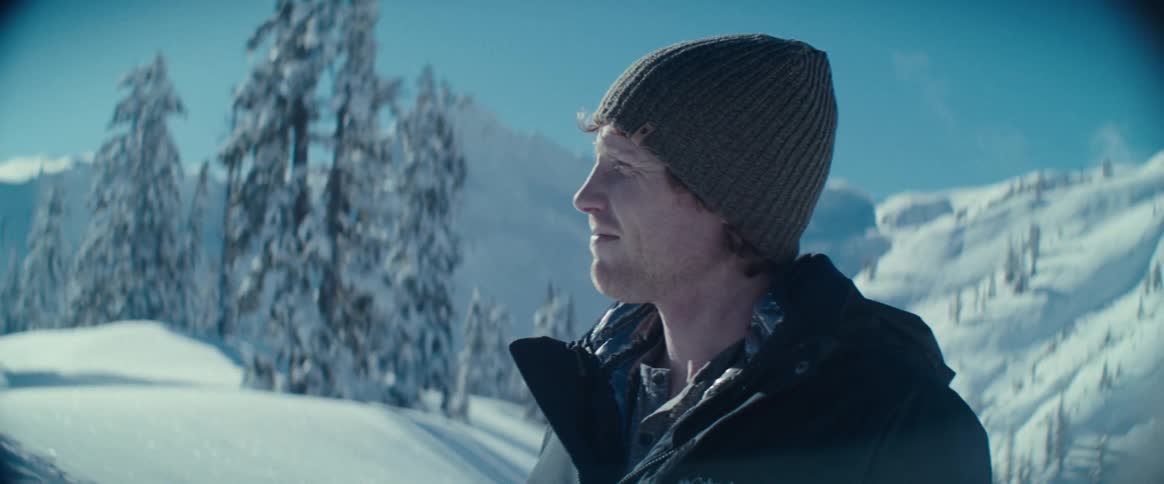 A man wearing a black jacket and a gray knit cap smiles while looking at a snowy landscape with sundappled pine trees and a mountain in the background