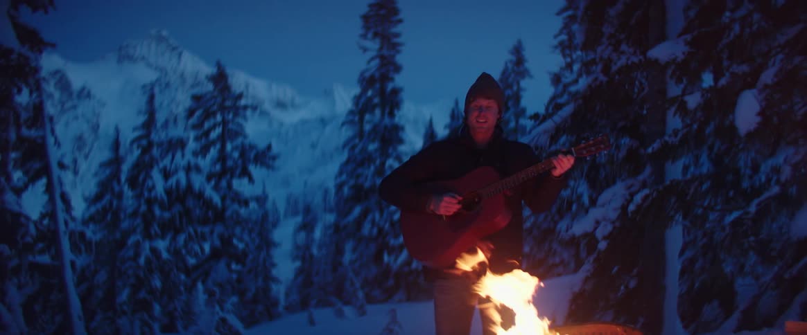 A man plays a guitar by a campfire in a snowy mountain forest at night