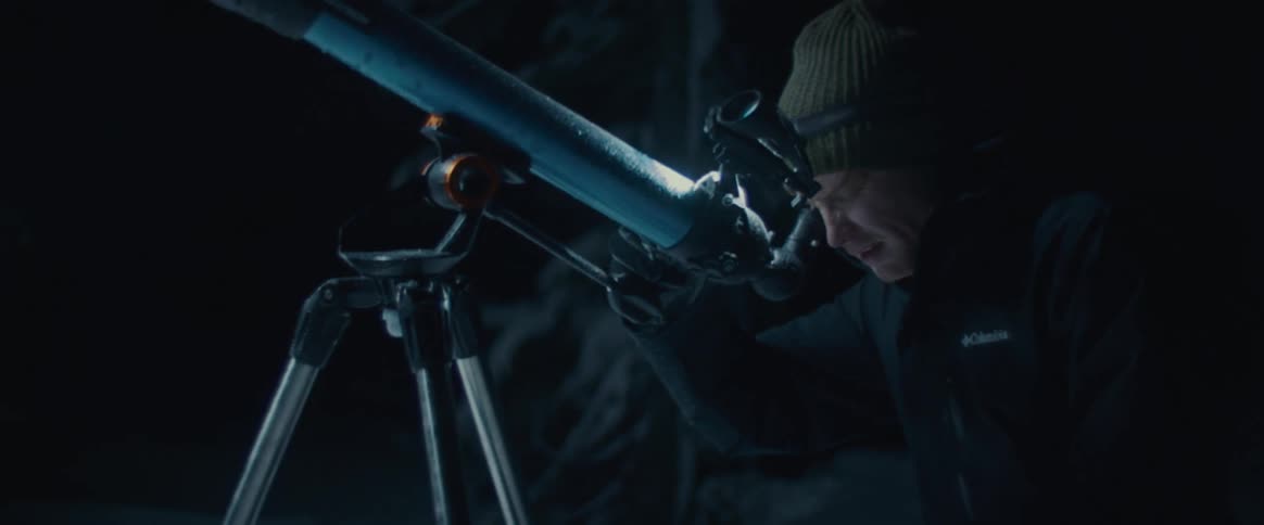 A man wearing a knit cap and a headlamp looks up from a telescope in the dark outdoors