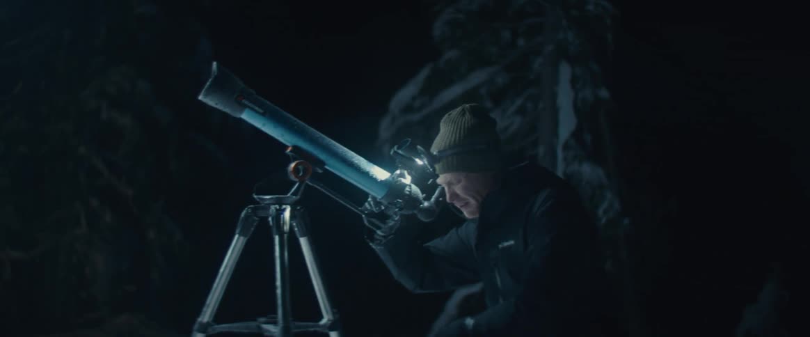 A man stargazes through a telescope in a snowy forest at night