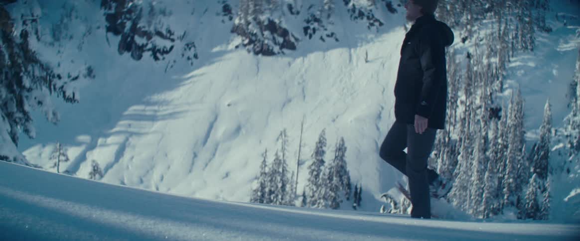 A man wearing a black jacket and a gray knit cap walks in deep snow on a white snowcovered mountain with large pine trees