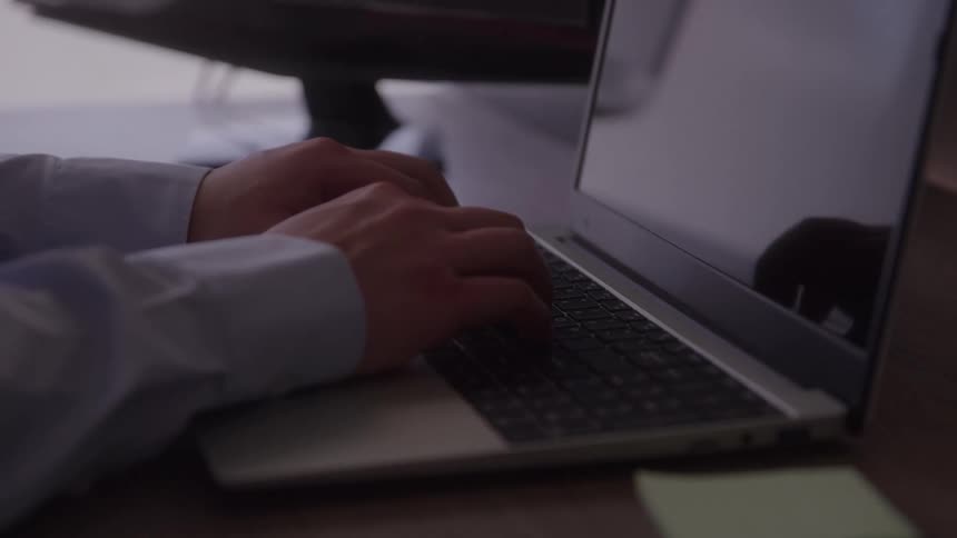 A close view shows a persons hands wearing a blue shirt as they type on a silver laptop keyboard on a dark desk with a sticky note