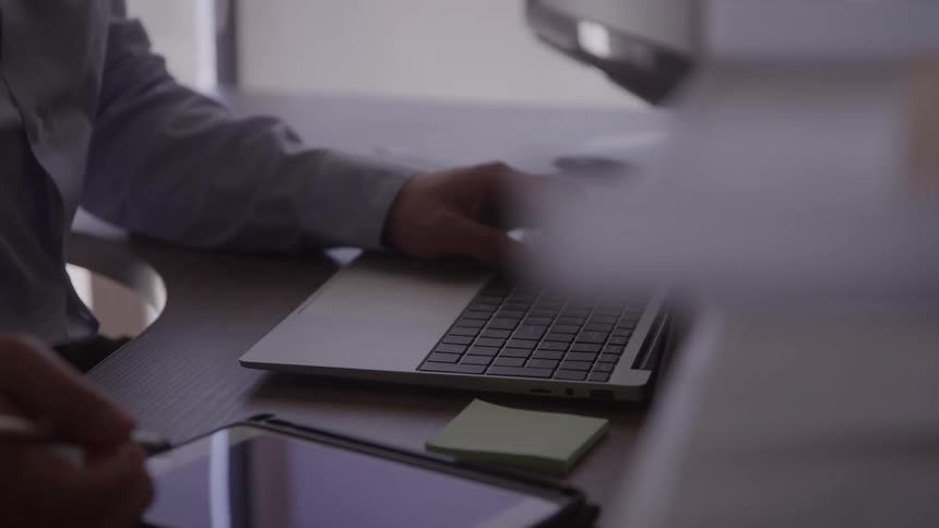 Man writing on a tablet with a pen at his desk right hand resting on a computer keyboard wearing a blue longsleeve shirt in his office at midday