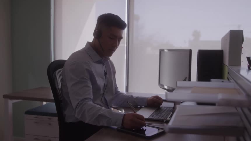 A young asian man using a tablet computer while sitting at a work desk in an office during daytime
