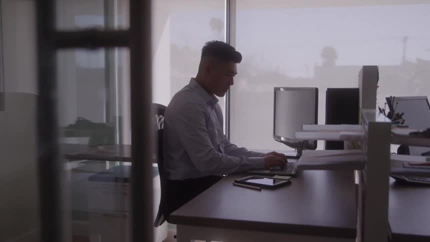 A man wearing a light blue dress shirt works on a laptop at a desk in a brightly lit office with large windows with filing cabinets and a computer monitor in the background