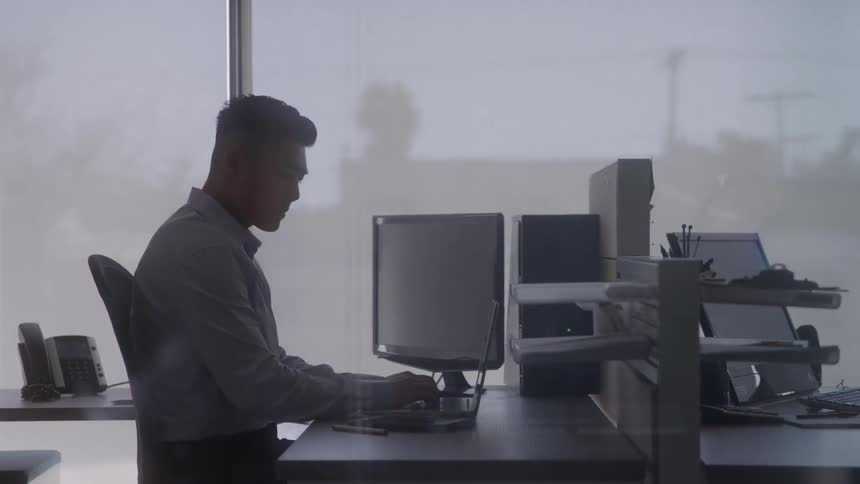 A male professional is sitting at a desk in an office facing away from the camera while working on a laptop the scene is brightly lit from the windows creating a silhouette effect that focuses on the individual and his task