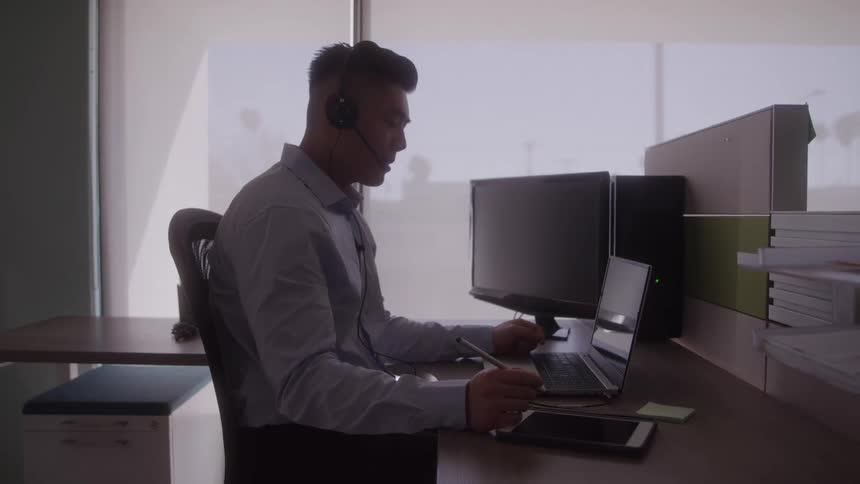 The video shows a man working at a desk in an office he is wearing a headset and appears to be in a virtual meeting or on a call actively using a pen on a digital tablet while looking at his laptop and a second monitor