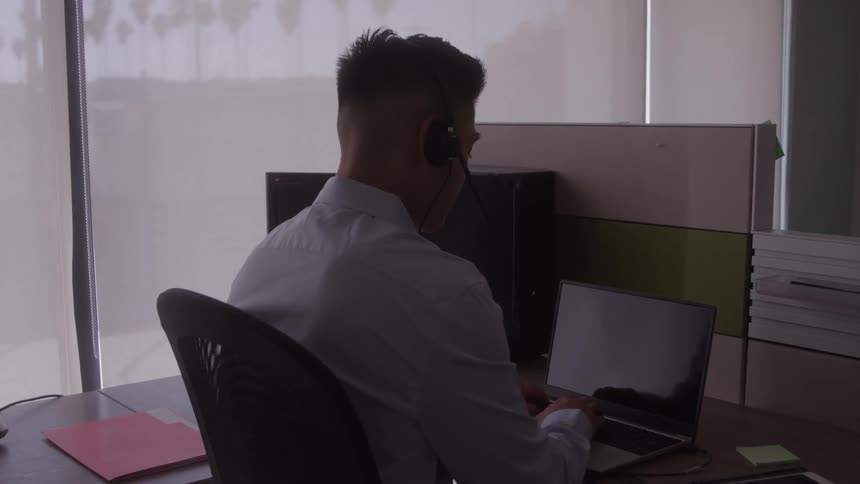 The video shows a man from behind sitting at a desk and working on a laptop he is wearing a headset and appears to be concentrating on his work in a professional office environment