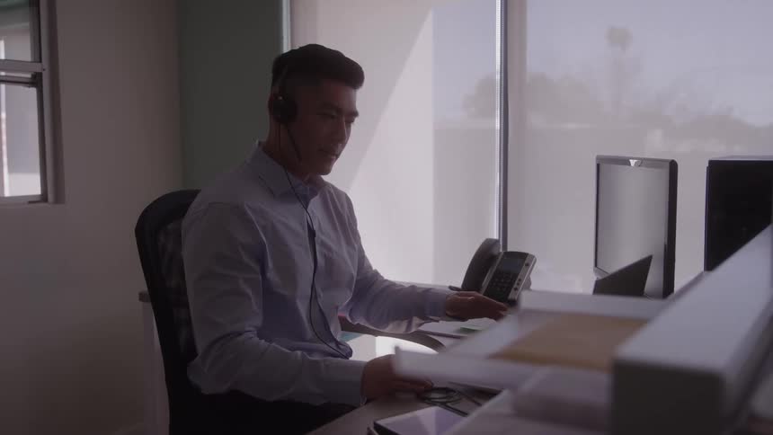 A person sits at a desk in an office while wearing a headset and looking at a computer while on a call