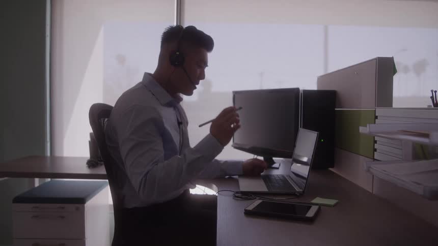 Young man wearing a longsleeve shirt and headset working behind a desk in his office at midday