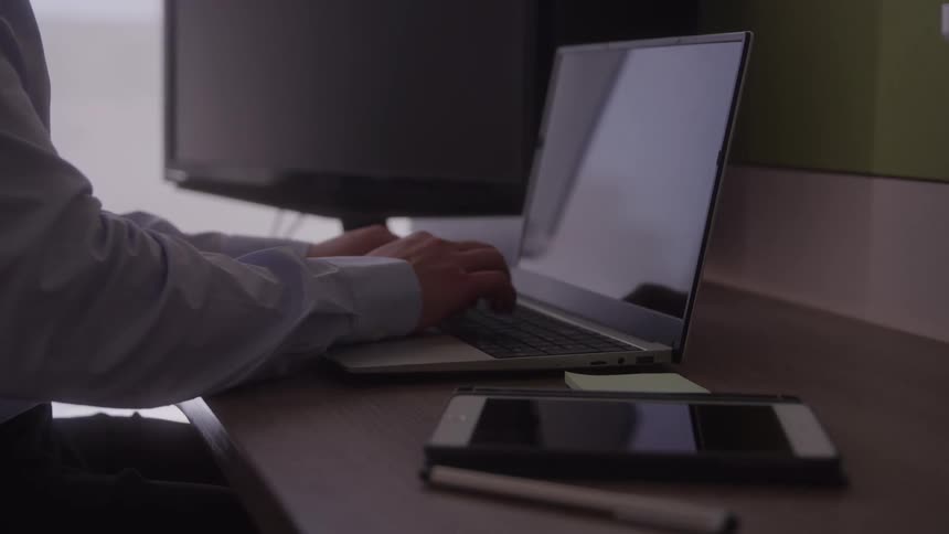 The video shows a closeup of a persons hands as they type on a laptop keyboard the person appears to be focused on their work with a smartphone and a digital pen on the desk beside the laptop