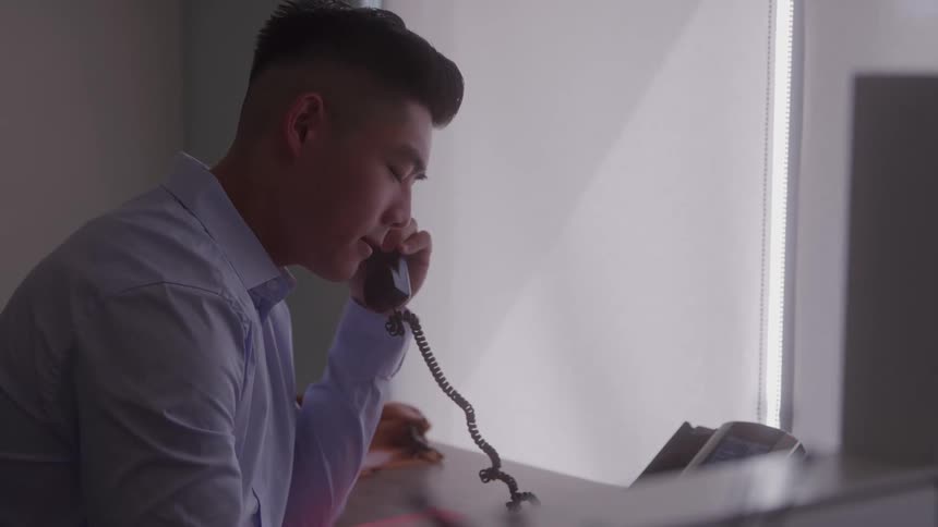 A young asian man is sitting at desk in an office answering a phone call and with a worried expression