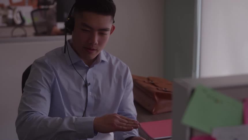 Young man wearing a headset sitting behind a computer at a desk inside an office at midday