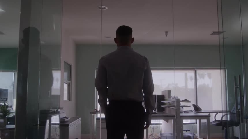 The video shows a man from behind standing in a dim office with his hands in his pockets he is standing still and looking out appearing to be contemplating something