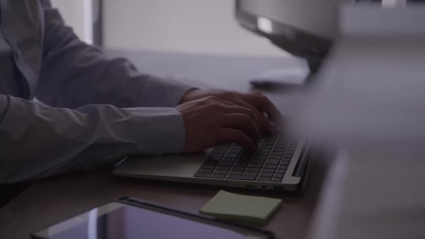 The video shows a mans hands as he types on a laptop keyboard he is working at a desk in an office with a tablet and a small notebook also visible nearby