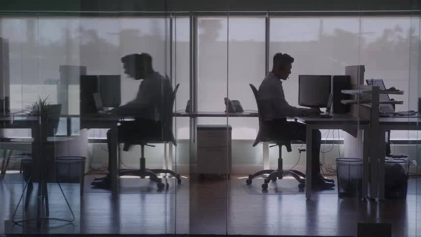 The video shows a wide shot of two men working at their desks in an office they are seated in front of computers with their silhouettes reflected in the glass walls of the workspace
