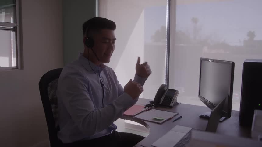 A young asian man is sitting at a desk in an office setting making hand gestures while answering a phone call
