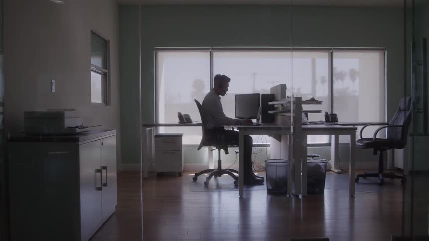 A man works alone on a laptop in a modern glass walled office