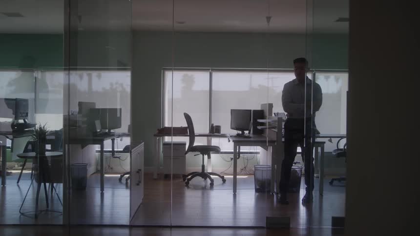 Young man walking left and right inside his office at midday deep in thought looking worried and sad rubbing his cheek with his right arm