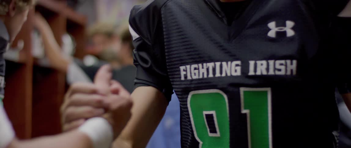A close view shows a football player in a black jersey with a green fighting irish logo shaking hands with a teammate in a locker room