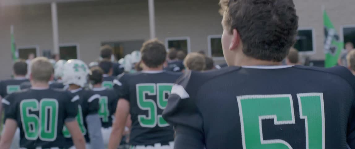 The video shows an american football team walking onto a field they are all wearing black and green jerseys with helmets with a focus on the back of a player whose jersey number is 51