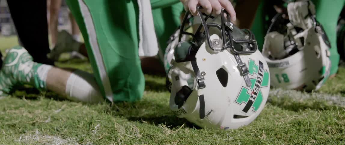 A close view shows a football player in a green and white uniform sitting on a green grass field holding his helmet with a fighting irish logo
