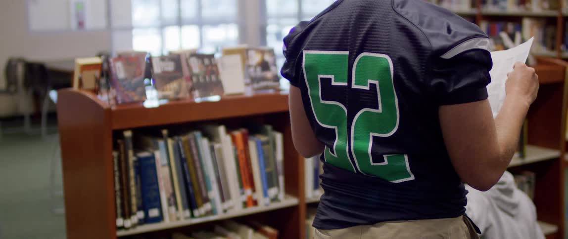 The video shows a young man wearing an american football jersey in a library he is standing in an aisle looking at the spines of the books on the shelves as if searching for a specific title