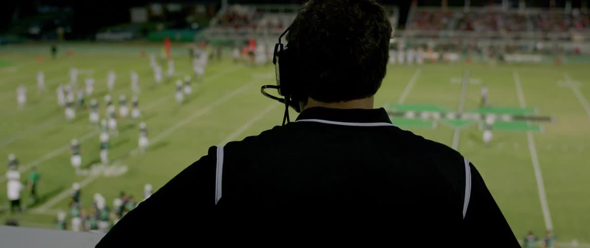 A coach wearing a headset watches a football game from above with the blurred players on the field below