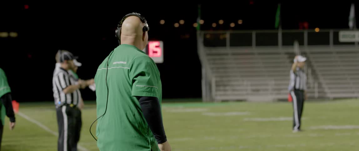 Referee inside a football pitch at night with another referee and a coaching official standing outside the pitch line watching the game