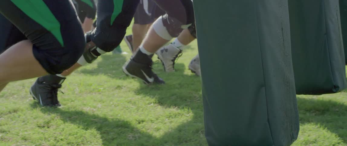 A close view shows a line of young football players in their gear with their feet and legs braced in a line on a green field during practice