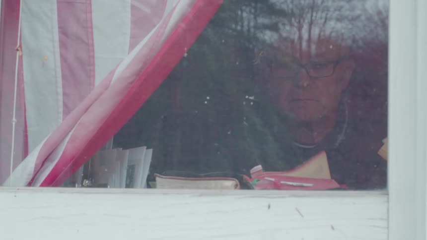 A shot through a window shows a man with glasses and a red and white flag possibly american draped to the left the mans face and shoulders are reflected in the window
