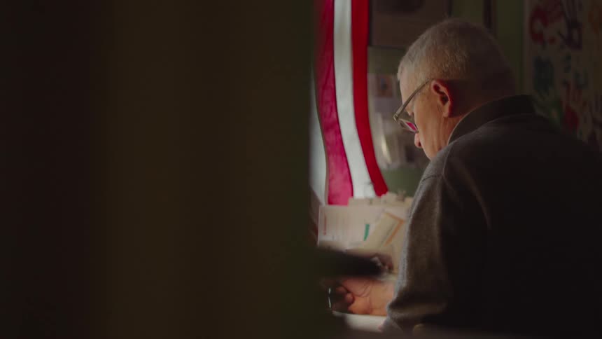 The video shows a side profile of a man sitting at a desk and writing the image is cropped in a way that two vertical lines are visible in the background a red line and a white line which appear to be part of an american flag hanging on the wall