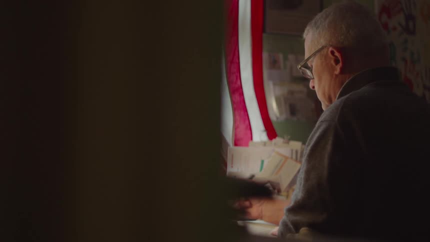 A side view of a man with gray hair and glasses is shown sitting at a table in a dimly lit room he is looking down at a book or papers in front of him and a red and white striped flag is partially visible in the background