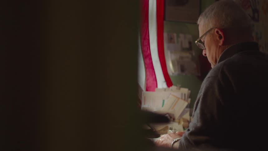 Man wearing glasses reads a book at a table with a flag in the background