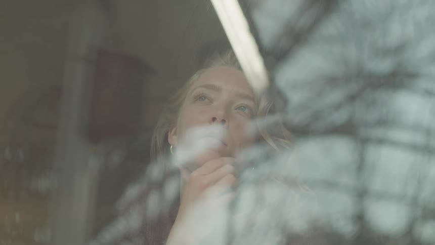 A young blonde woman is looking through the window with a contemplative expression