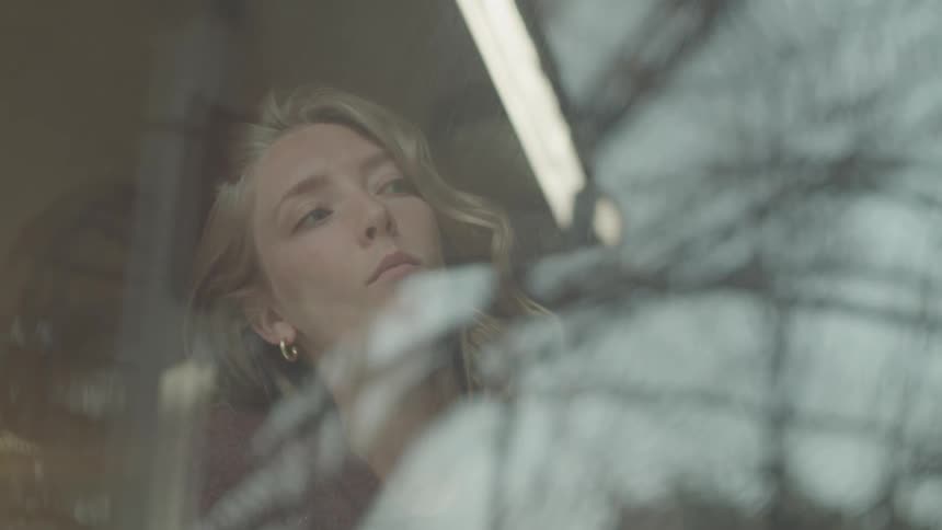 A person with lightcolored hair and a reflective gaze looks out of a window the shot is captured through the glass which creates a hazy reflective effect