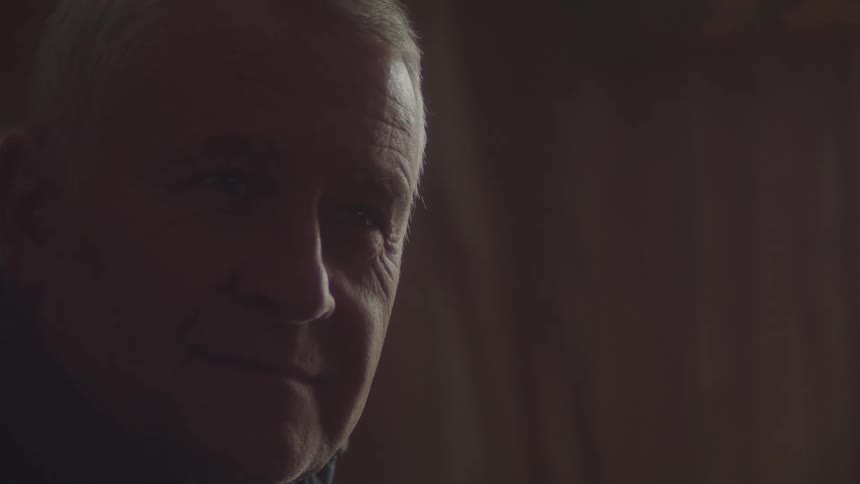 A lowangle closeup shot of an older mans face with the left side in shadow and the right side partially illuminated the background is a dark wooden wall