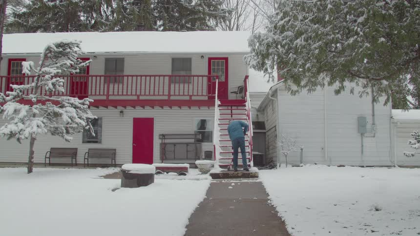 A man is in front of a house with red doors shoveling snow from the stairs